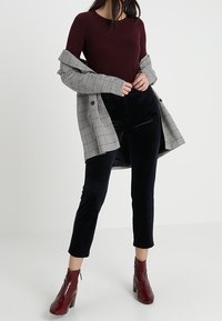 Bordová dlouhá halenka vrstvená pod šedým károvaným oversized kabátem, s černými sametovými kalhotami po kotníky a lesklými burgundskými botami na podpatku.