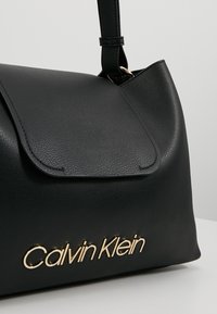 Sac à main en cuir noir avec rabat et logo Calvin Klein doré sur le devant, doté d'une bandoulière unique attachée par un anneau métallique.