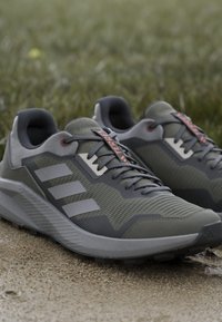 Baskets de sport grises avec un dessus texturé, trois bandes horizontales et une semelle extérieure robuste. Des lacets avec des accents orange ajoutent une touche de détail.
