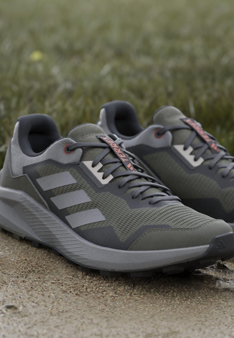 Baskets de sport grises avec un dessus texturé, trois bandes horizontales et une semelle extérieure robuste. Des lacets avec des accents orange ajoutent une touche de détail.