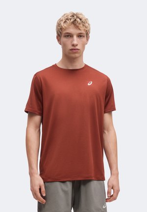 Homme blond portant un t-shirt de sport à manches courtes couleur rouille et un short gris, debout face à un fond uni.
