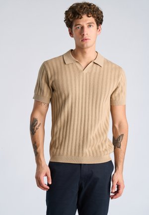 Junger Mann mit lockigem Haar, der ein beigefarbenes kurzärmliges Strick-Poloshirt und dunkle Hosen trägt, mit sichtbaren Tätowierungen auf beiden Unterarmen.