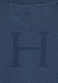 Tamno plava tkanina s velikim utisnutim slovom "H" i manjim utisnutim tekstom "HACKETT" blizu izreza.