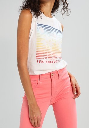 Vrouw draagt een mouwloos wit Levi's top met een kleurrijke print van zonsondergang en palmbomen, gecombineerd met koraalroze strakke jeans.