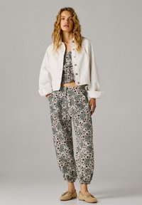 Kobieta ubrana w białą zapinaną na guziki kurtkę, pod którą ma kwiecisty crop top i luźne spodnie w tym samym wzorze, na nogach beżowe mokasyny, stoi na tle jednolitego tła.