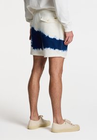 Shorts de tie-dye blanco y navy que presentan una textura suave, cinturilla elástica y un bolsillo trasero con cierre a botón, combinados con zapatos claros.