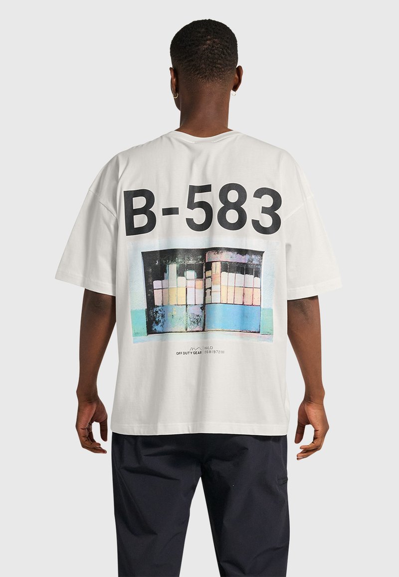 Vit oversize t-shirt med en grafisk design med "B-583" och en mönstrad bakgrund av färgglada rektanglar, tillverkad av bomull.