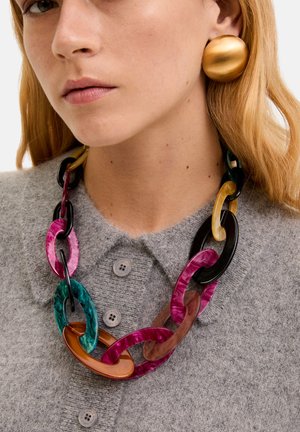 Kleurrijke acrylaatketting met oversized schakels in groen, roze, zwart en bruin, gecombineerd met een gestructureerde grijze trui en gouden oorbellen.