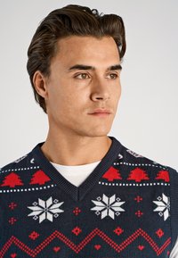 Suéter de cuello en V azul marino con patrones festivos rojos y blancos, incluyendo árboles y copos de nieve. Hecho de tela de punto con una textura suave.