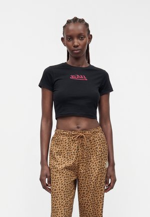 Femme aux cheveux tressés portant un t-shirt court noir Von Dutch et un pantalon à cordon avec motif léopard, debout sur un fond blanc uni.