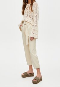 Pull crème en maille dentelle avec manches évasées, noué au niveau du col, associé à un pantalon beige ceinturé et des sandales plateformes tissées.