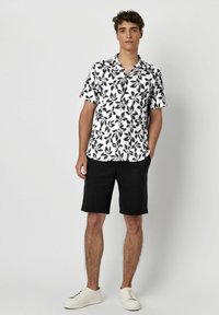 Kurzarmhemd in Schwarz und Weiß mit Blattmuster. Getragen mit schwarzen Shorts und weißen Sneakers. Minimalistisches Design mit lockerer Passform.