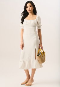 White dress with puffed sleeves, square neckline, side slit, and ruffled hem. Model holds a woven bag with yellow handles and wears yellow flip-flops.