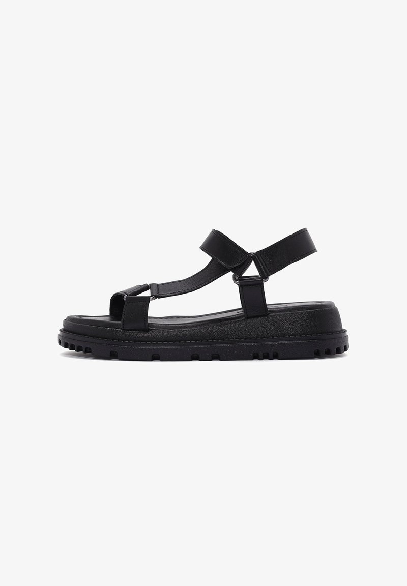 Black sandal with a flat sole, featuring a textured finish, adjustable straps, and a triangular hardware accent for added detail.