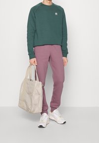 Grön bomullströja med vit logo, lila sweatshirtbyxor, beige tygväska och vita sneakers med gröna detaljer.