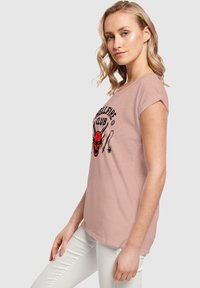 Camiseta de manga corta rosa claro con un diseño gráfico en negro y rojo que presenta motivos de diablo y estrellas. Fabricada en un material suave y transpirable.