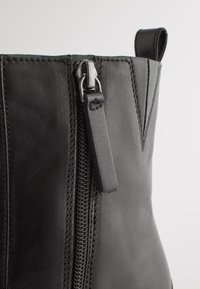 Bottine en cuir noir avec une fermeture éclair argentée et des détails de couture. Texture lisse avec une languette pour un enfilage facile.
