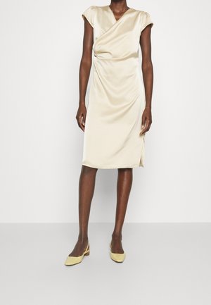 Femme portant une robe en satin crème longueur genou avec des manches courtes et des chaussures slingback jaunes à petit talon sur un fond uni.