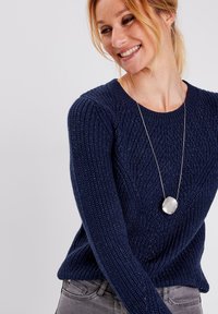 Marineblauer gestrickter Pullover mit einem strukturierten Muster, Rundhalsausschnitt und langen Ärmeln. Kombiniert mit einer silbernen Anhängerkette.