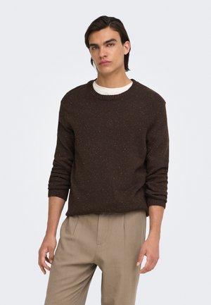 Jeune homme aux cheveux foncés portant un pull marron foncé moucheté par-dessus une chemise blanche et un pantalon beige, debout devant un fond clair uni.