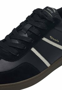 Baskets noires en cuir et daim, avec des lacets noirs, deux rayures blanches et une semelle en caoutchouc marron. Nom de la marque "Tamaris" affiché en doré.