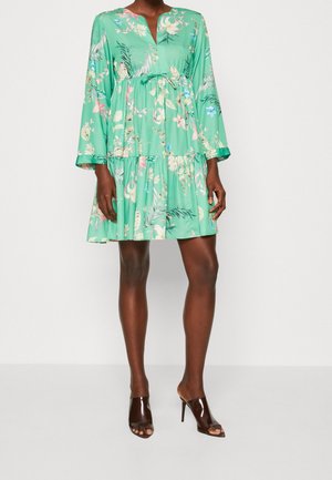 Femme portant une robe verte courte et ample à manches longues avec un imprimé floral, associée à des chaussures à talons hauts ouvertes marron foncé.