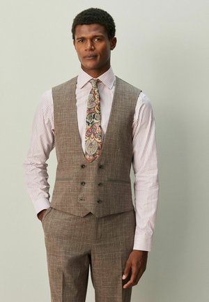 Muž oblečený v hnedom kockovanom oblekovom veste a nohaviciach, ružovej pruhovanej košeli a kravate s motívom paisley, stojaci s jednou rukou v vrecku.