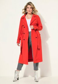 Rode trenchcoat met zwarte knopen en taillekoord, gecombineerd met grijze jeans en metallic zilveren enkellaarsjes, tegen een neutrale achtergrond.