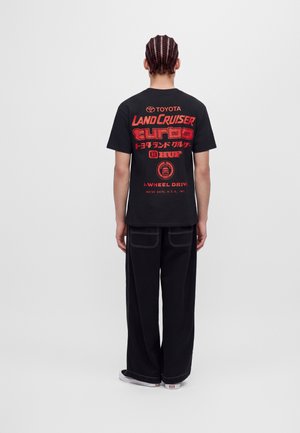 Un modèle aux cheveux tressés est de dos et porte un T-shirt noir avec un motif Toyota Land Cruiser, un pantalon noir ample et des baskets, sur un fond uni.