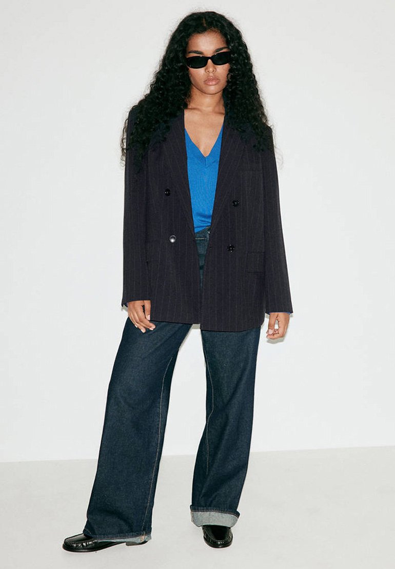 Femme aux longs cheveux bouclés portant des lunettes de soleil foncées, un blazer noir à fines rayures, un haut bleu à col en V, un jean large et des chaussures noires, debout contre un mur blanc.
