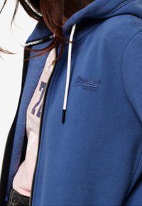 Blue hooded sweatshirt with white drawstrings, soft texture, zip closure, and embossed logo on the chest. Worn over a pink graphic t-shirt.