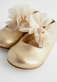 Guld läderskor med en texturerad yta, prydda med stora beige tyllblommor på framsidan, rundade tå och platta sulor.