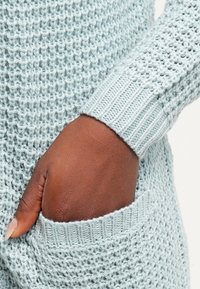 Jersey de lana azul claro con un patrón texturizado, con puños acanalados y un bolsillo delantero. La tela parece suave y transpirable.