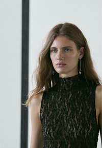 Zwarte kanten jurk met een hoge kraag, met een gestructureerd patroon, gedeeltelijk transparant ontwerp, en lange oorbellen met parels.