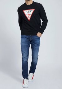 Sudadera negra con un logo triangular en blanco y rojo; jeans azules; zapatillas blancas con detalles rojos; exhibición de un atuendo casual.