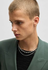 Grüner Blazer mit Spitzenrevers, getragen über einem strukturierten schwarzen Pullover. Zwei silberne Halsketten, eine mit runden Perlen, prominent geschichtet.
