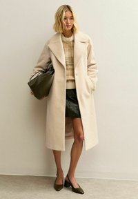 Mujer vestida con abrigo largo beige, suéter de patrones, falda negra texturizada, zapatos de tacón verdes y llevando un bolso grande verde, de pie contra una pared lisa.