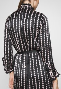 Vestido de satén negro con un patrón vertical de puntos rosas, cuello con volantes, cierres de botones y puños de manga fruncidos. Diseñado con un cinturón en la cintura.