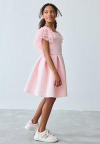 Tyttö, jolla on kiharat hiukset ja vaaleanpunainen mekko, jossa on kukalliset puffy hihat, sekä valko-vaaleanpunaiset lenkkarit, seisoo harmaalla lattialla valkoista seinää vasten.