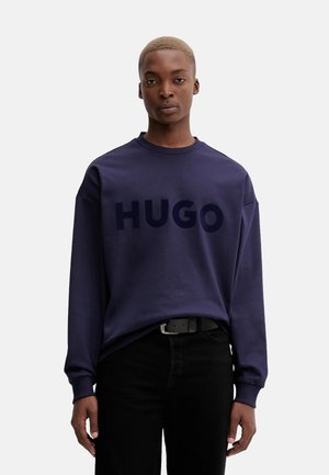Sudadera azul marino con un atrevido logo "HUGO" en azul oscuro, fabricada en un tejido suave, con cuello redondo y un corte relajado. Jeans negros y cinturón visibles.