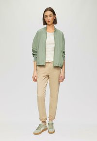 Světle zelená bomber bunda, béžový pruhovaný top, béžové chino kalhoty a světle zelené tenisky s gumovou podrážkou. Model stojí uprostřed s neutrálním pozadím.