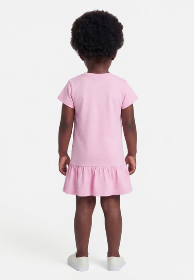 Παιδί με σκουρόχρωμο δέρμα και σγουρά μαλλιά, φορώντας ένα ροζ φόρεμα με κοντά μανίκια και λευκά παπούτσια, στέκεται γυρισμένο με την πλάτη προς τα εμπρός σε λευκό φόντο.