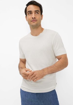 Mann trägt ein weißes kurzärmliges T-Shirt und blaue Hose, schaut leicht nach oben und zur Seite, die Hände locker verschränkt.