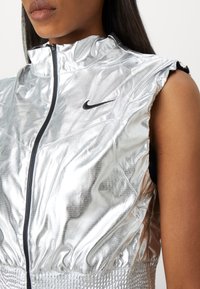 Silberne Nike ärmellose Jacke mit hohem Kragen, ausgestattet mit einem Reißverschluss auf der Vorderseite und strukturiertem Stoff, akzentuiert durch ein schwarzes Logo auf der Brust.