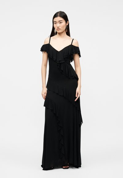 Γυναίκα που φοράει μαύρο μάξι φόρεμα με ανοιχτούς ώμους και διαγώνιες πιέτες, στέκεται με τα χέρια χαλαρά, μπροστά σε απλό ανοιχτόχρωμο φόντο.