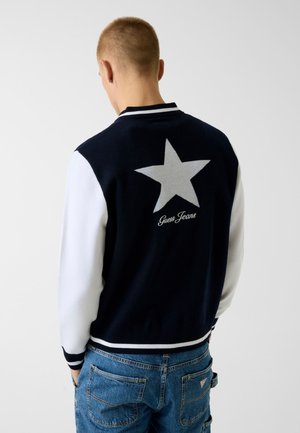 Osoba odwrócona tyłem, ubrana w kurtkę typu varsity z białymi rękawami, dużą gwiazdą i napisem "Guess Jeans" na plecach oraz niebieskie dżinsy.
