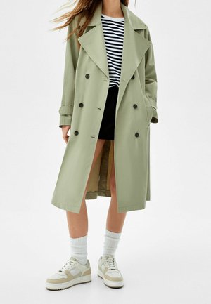 Person iført en lysegrøn dobbeltradet trenchcoat, sort nederdel, stribet skjorte, hvide sokker og beige-hvide sneakers, stående på hvid baggrund.