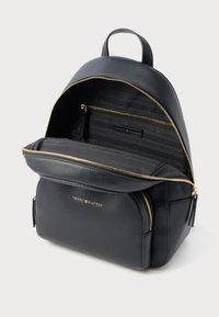Černý kožený batoh se zlatým zipem, přední kapsou a embosovanou vnitřní podšívkou s logem. Kompaktní kulatý design s horním uchycením.