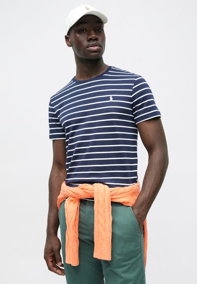 Μπλε ναυτικό μπλουζάκι με λευκές ρίγες, με μικρό λογότυπο. Φορεμένο με πράσινο παντελόνι και πορτοκαλί πουλόβερ με πλέξη καλώδιο, δεμένο στη μέση.