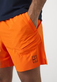Homme portant un short de sport orange vif avec une poche latérale zippée et un logo Nike noir sur la jambe gauche, la main reposant dans la poche avant.
